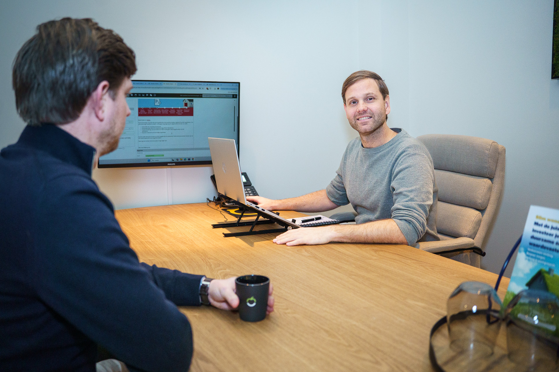 2 mannen zitten aan een bureau, 1 kijkt de camera in