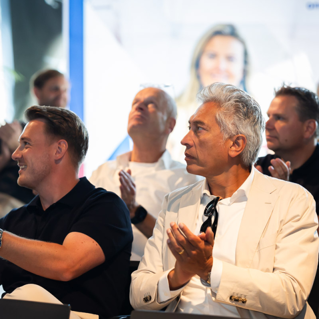 Mannen zitten op stoelen naast elkaar en klappen